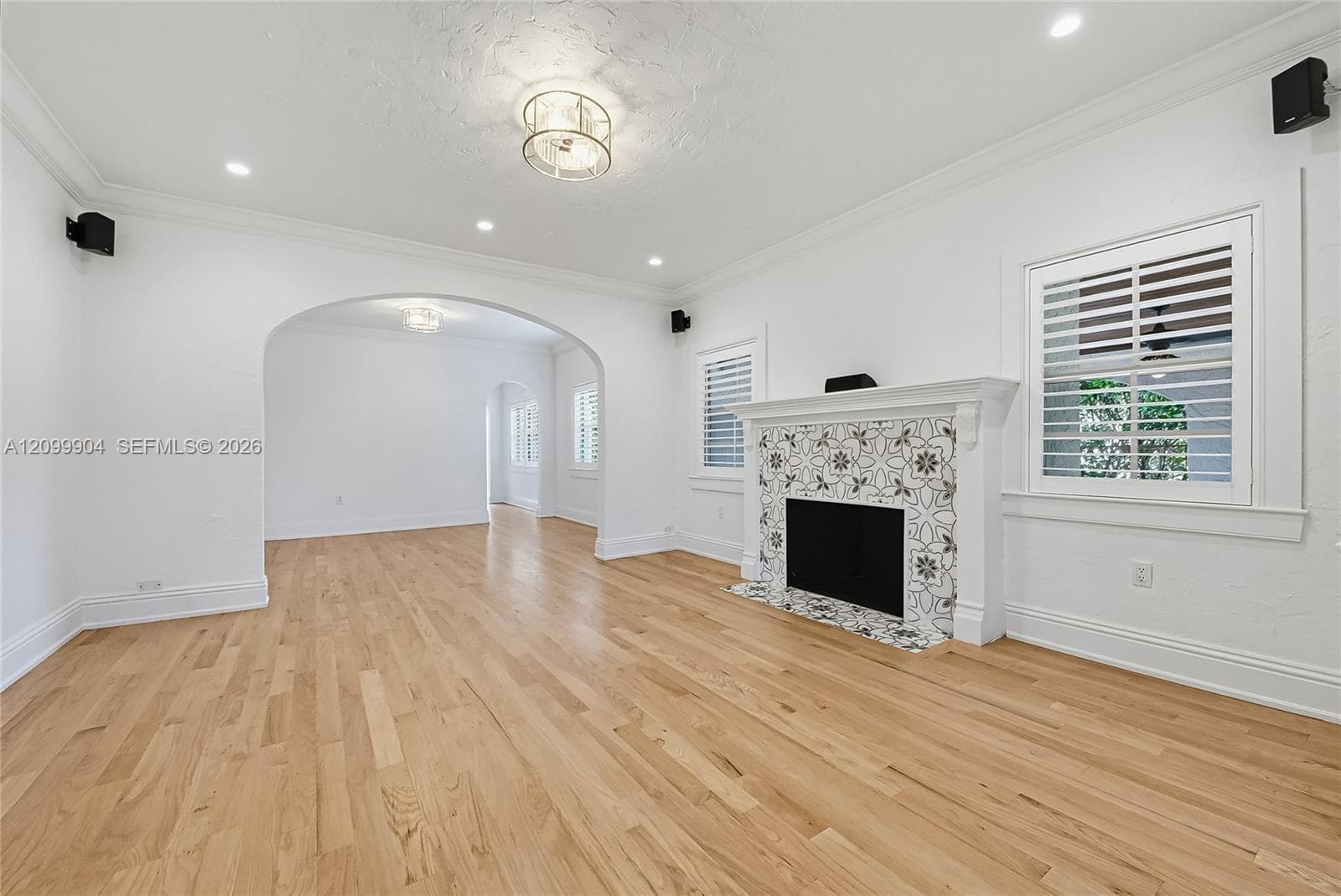 Living area featuring wood-finish flooring, a decorative tile-surround fireplace, arched architectural entryways, crown molding, and plantation shutters
