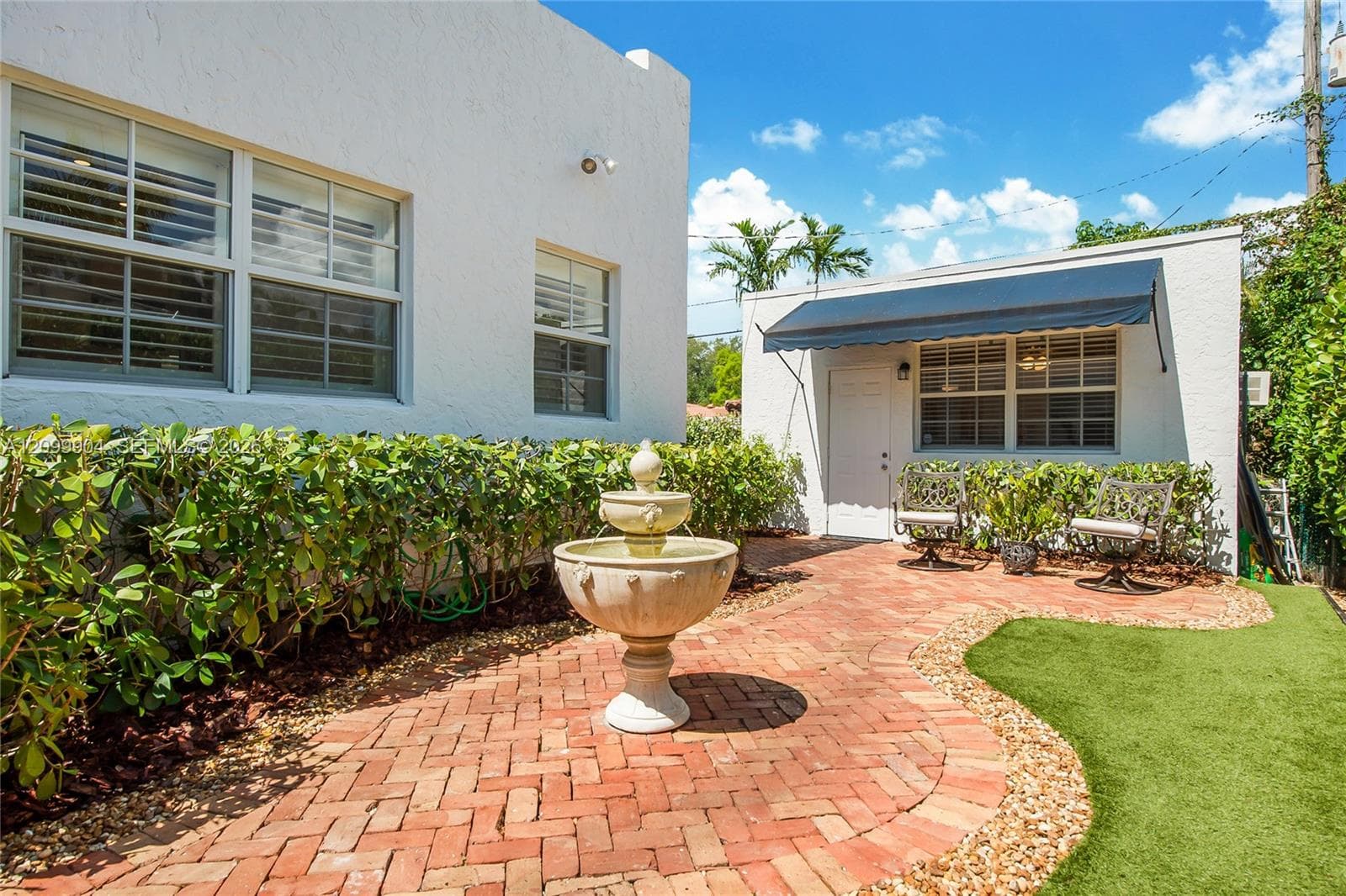 Courtyard featuring a herringbone-pattern brick patio, a central tiered stone fountain, exterior building structures with textured white stucco siding, and a retractable dark-toned window awning