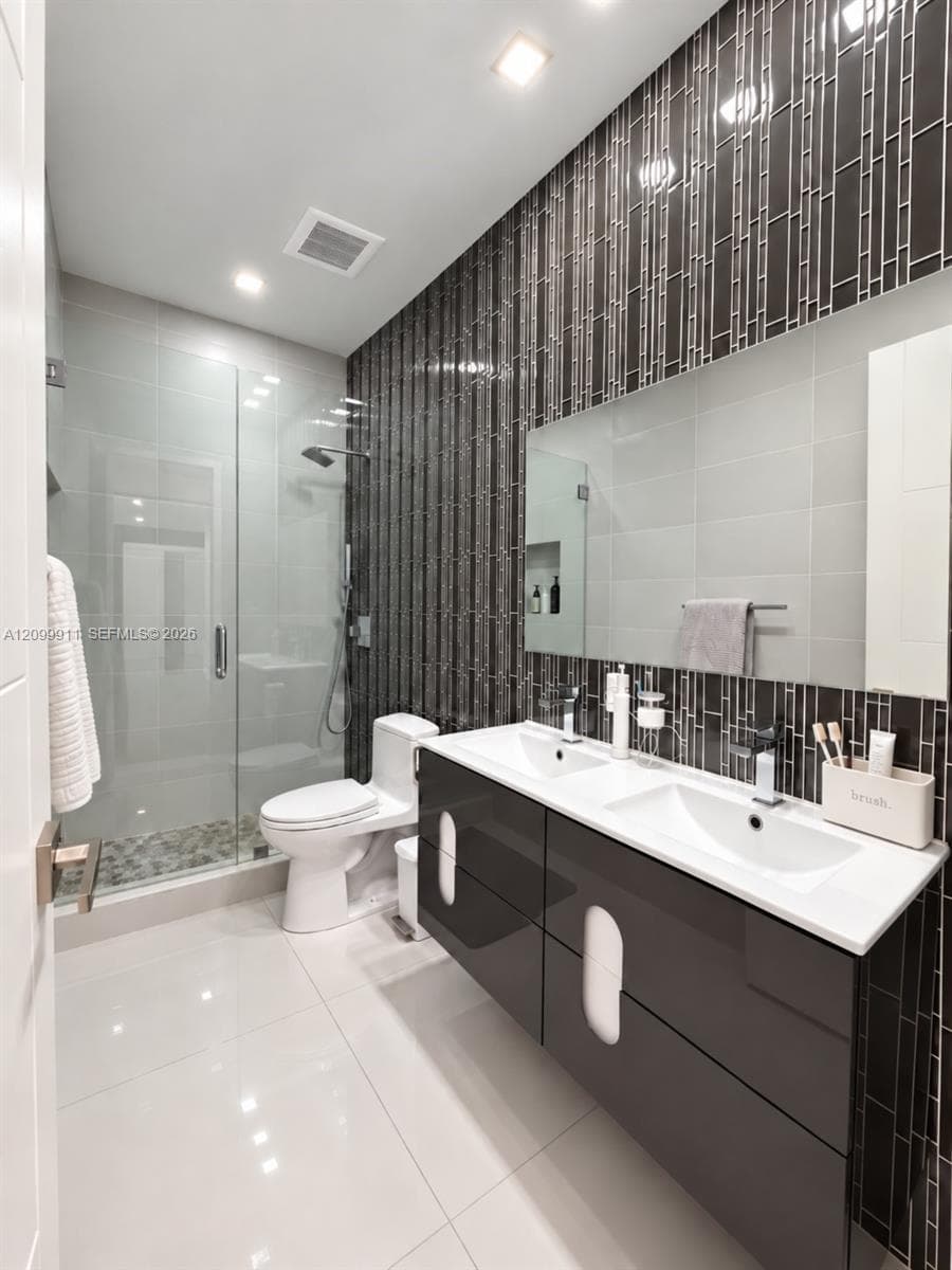 Floating double vanity with a composite countertop and integrated dual sinks, vertical textured tile wall detail, frameless glass-enclosed shower with a pebble-stone floor, and large-format porcelain floor tiling