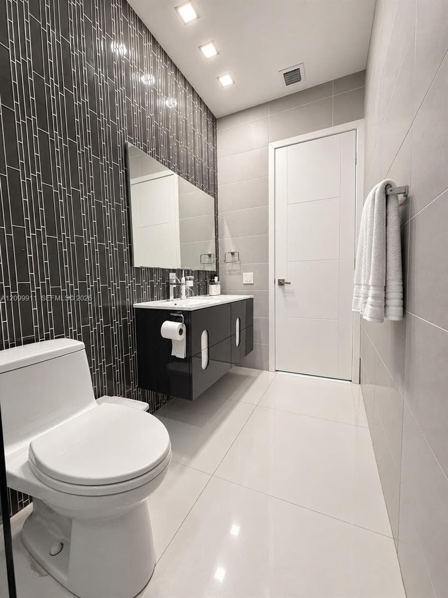 Bathroom featuring large-format white tile flooring, wall-mounted black cabinetry, a vertical-patterned dark tile accent wall, recessed lighting, and a frameless mirror