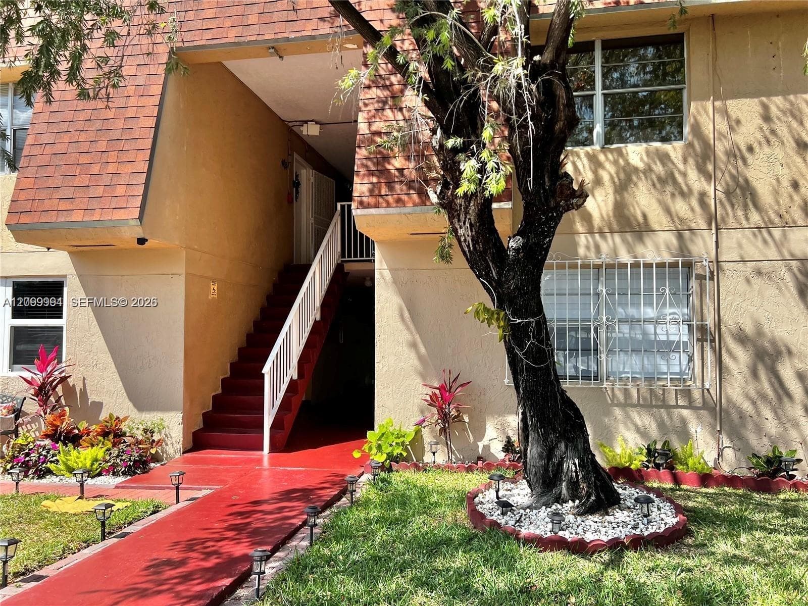 Multi-level building exterior featuring a red tile roof, red-painted concrete walkway, white-painted exterior staircase, and mature landscaping
