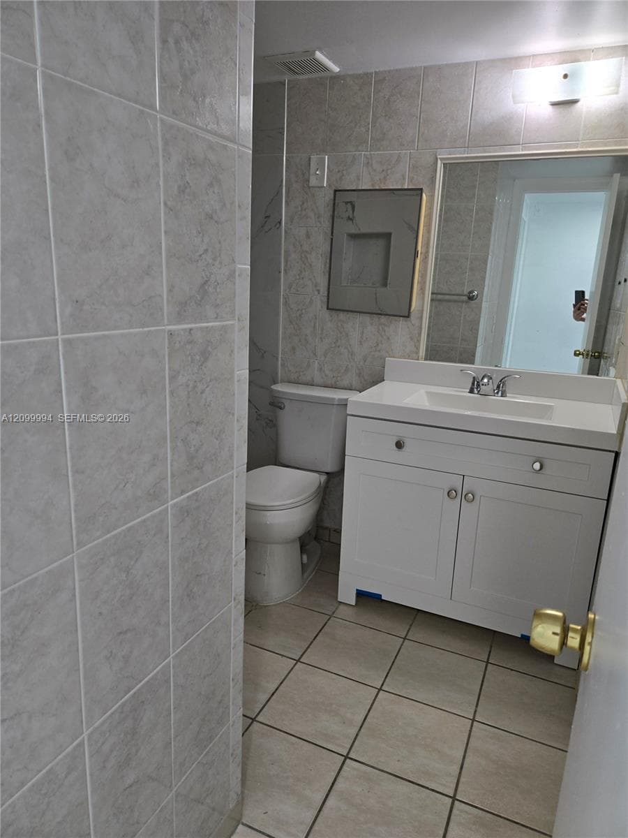 Bathroom featuring a white vanity cabinet with a stone-top integrated sink, neutral wall-to-wall ceramic tile, recessed shower niche, and a wall-mounted rectangular mirror