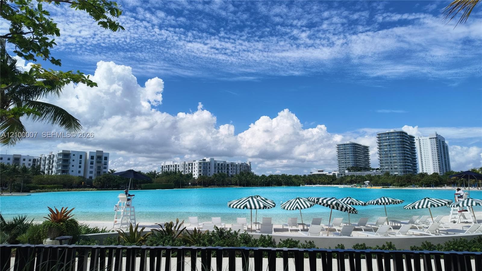 Expansive water feature with white sand shore, multiple striped umbrellas, and mature landscaping