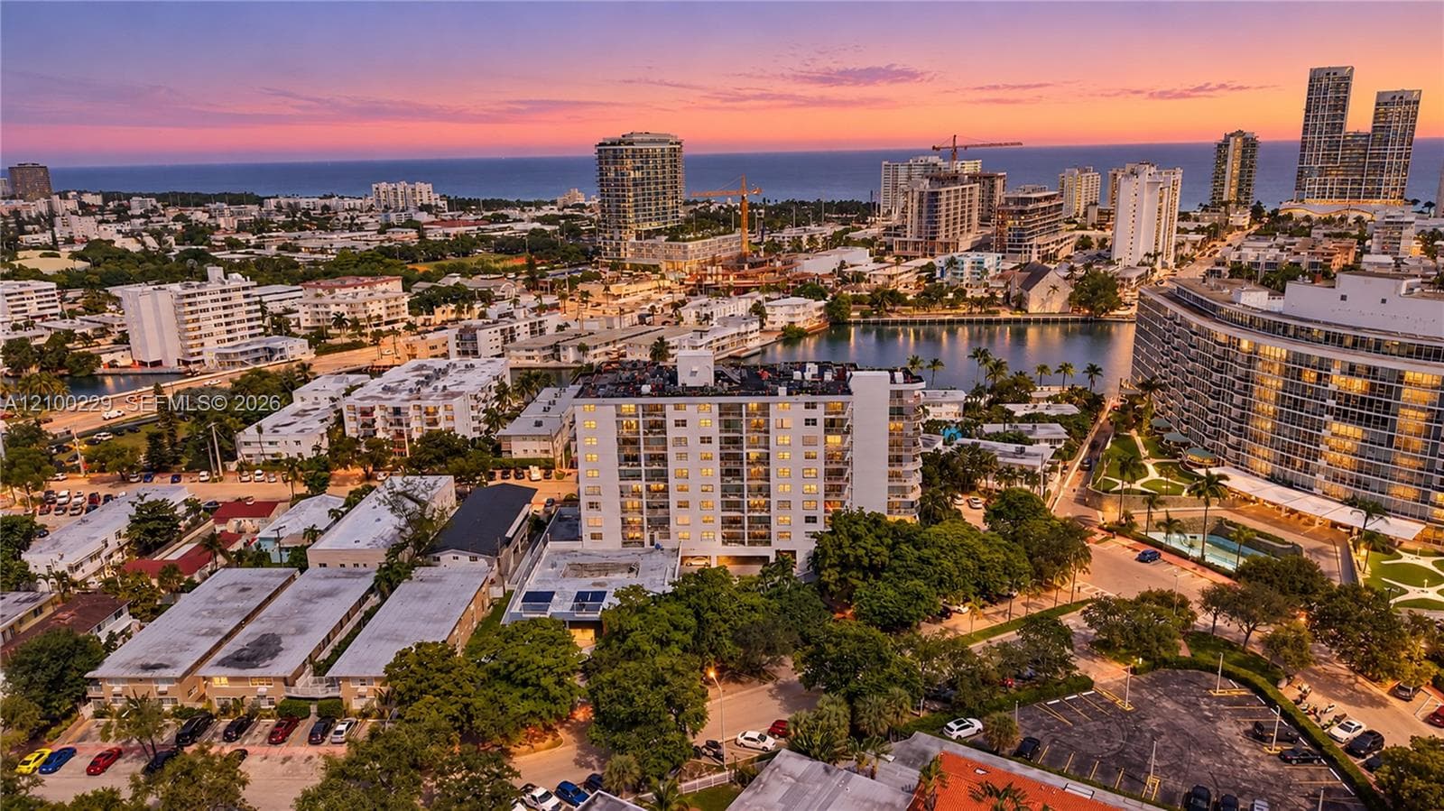 Aerial perspective of a high-rise residential building featuring a multi-story structure with private balconies, surrounded by mature trees, paved parking areas, and proximity to local waterways and urban coastline