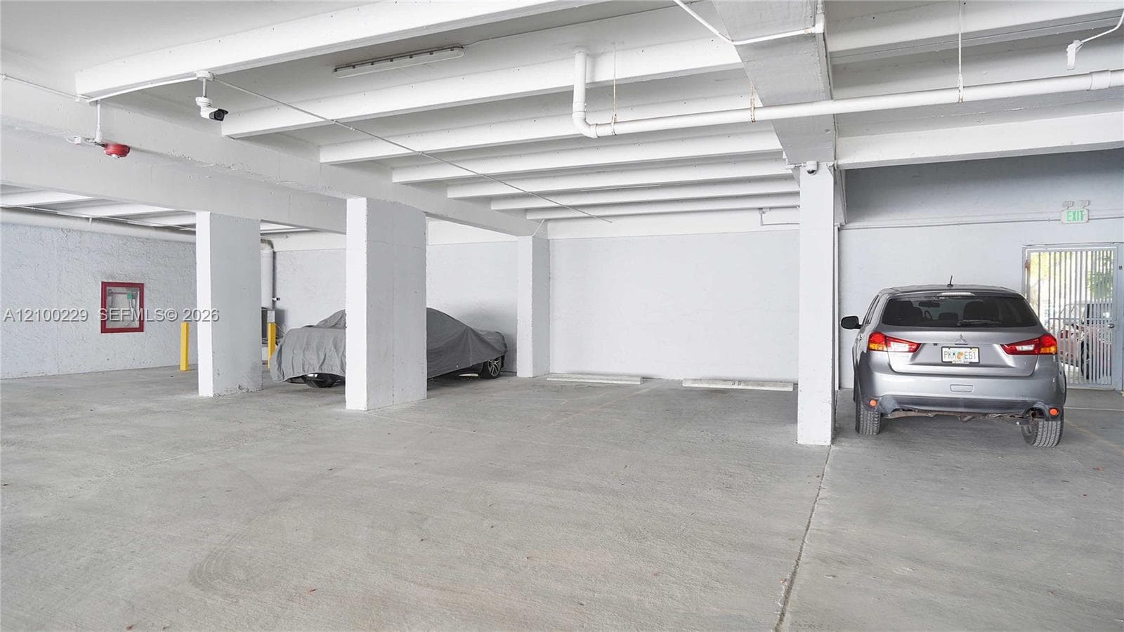 Covered parking area featuring structural concrete columns, a painted concrete floor, exposed ceiling joists, and security lighting