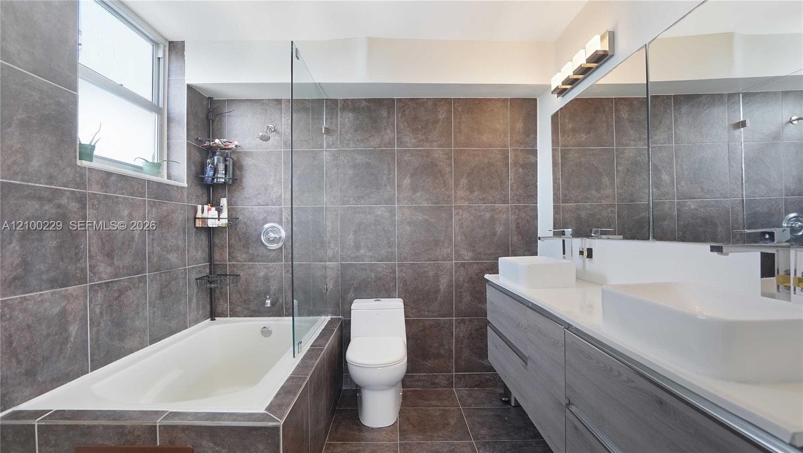 Bathroom featuring large-format stone-finish wall and floor tile, a glass-enclosed tub, dual vessel sinks, a wood-finish vanity, and full-width vanity mirror