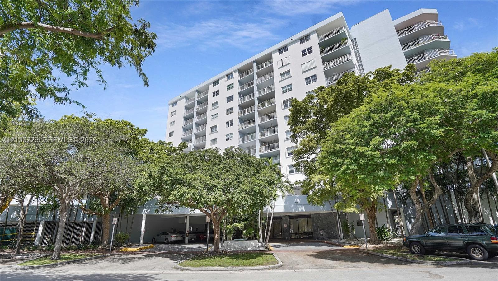 High-rise building exterior featuring private balconies, integrated ground-level covered parking, and mature surrounding landscaping