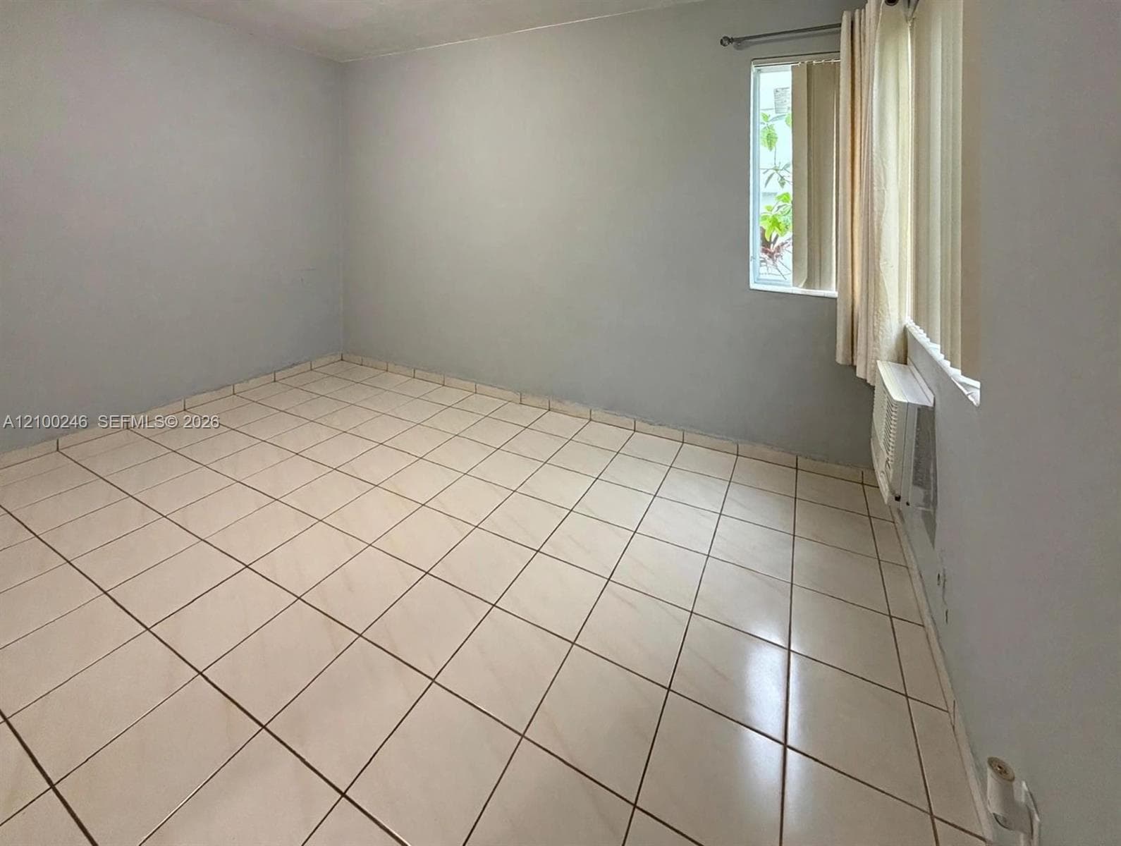 Neutral-toned interior space featuring light-colored square floor tiling, solid-colored walls, a window with vertical blinds, and a wall-mounted air conditioning unit