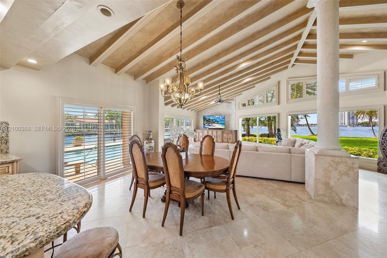 Open-concept living space featuring vaulted ceilings with exposed wood beams, natural stone flooring, a decorative stone column, and expansive floor-to-ceiling windows with waterfront exposure