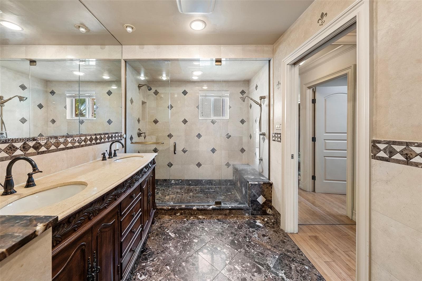 Dual-sink vanity with ornate wood cabinetry, stone countertops, walk-in glass shower enclosure with built-in bench, and patterned stone tile flooring