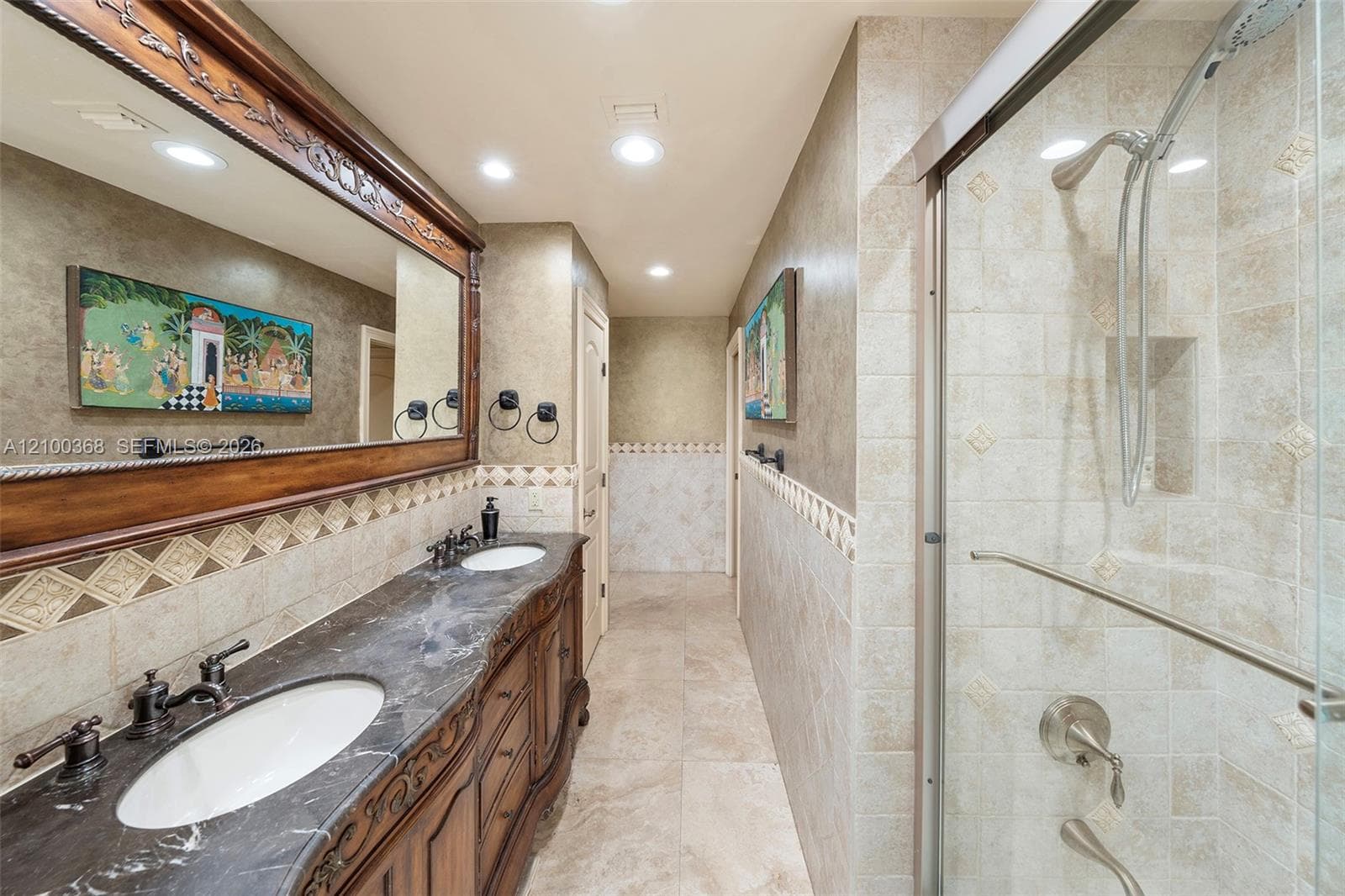 Double vanity with stone countertop, dual undermount sinks, intricate wood-finish cabinetry, a large framed vanity mirror, and a tiled walk-in shower with glass enclosure