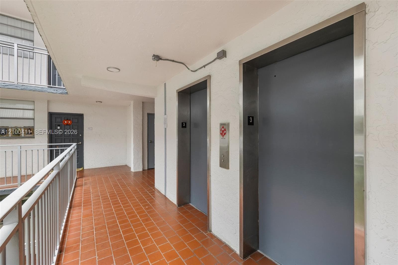 Covered walkway featuring terracotta tile flooring, metal railings, and access to dual elevators with stainless steel frames