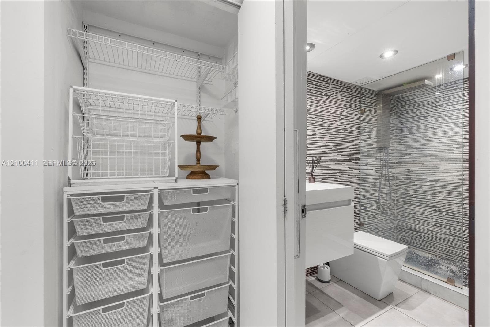 Walk-in closet featuring wire shelving and a modular storage system, paired with a bathroom containing a wall-mounted vanity, square-profile toilet, glass-enclosed shower with horizontal textured tile, and recessed lighting