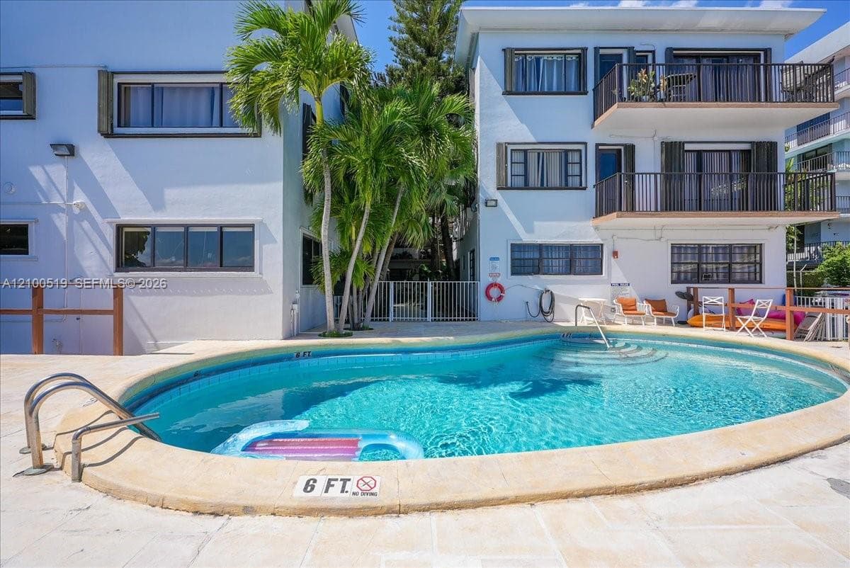 Oval swimming pool with integrated ladder and stone-finish coping, multi-level structure with private balconies, and mature palm trees