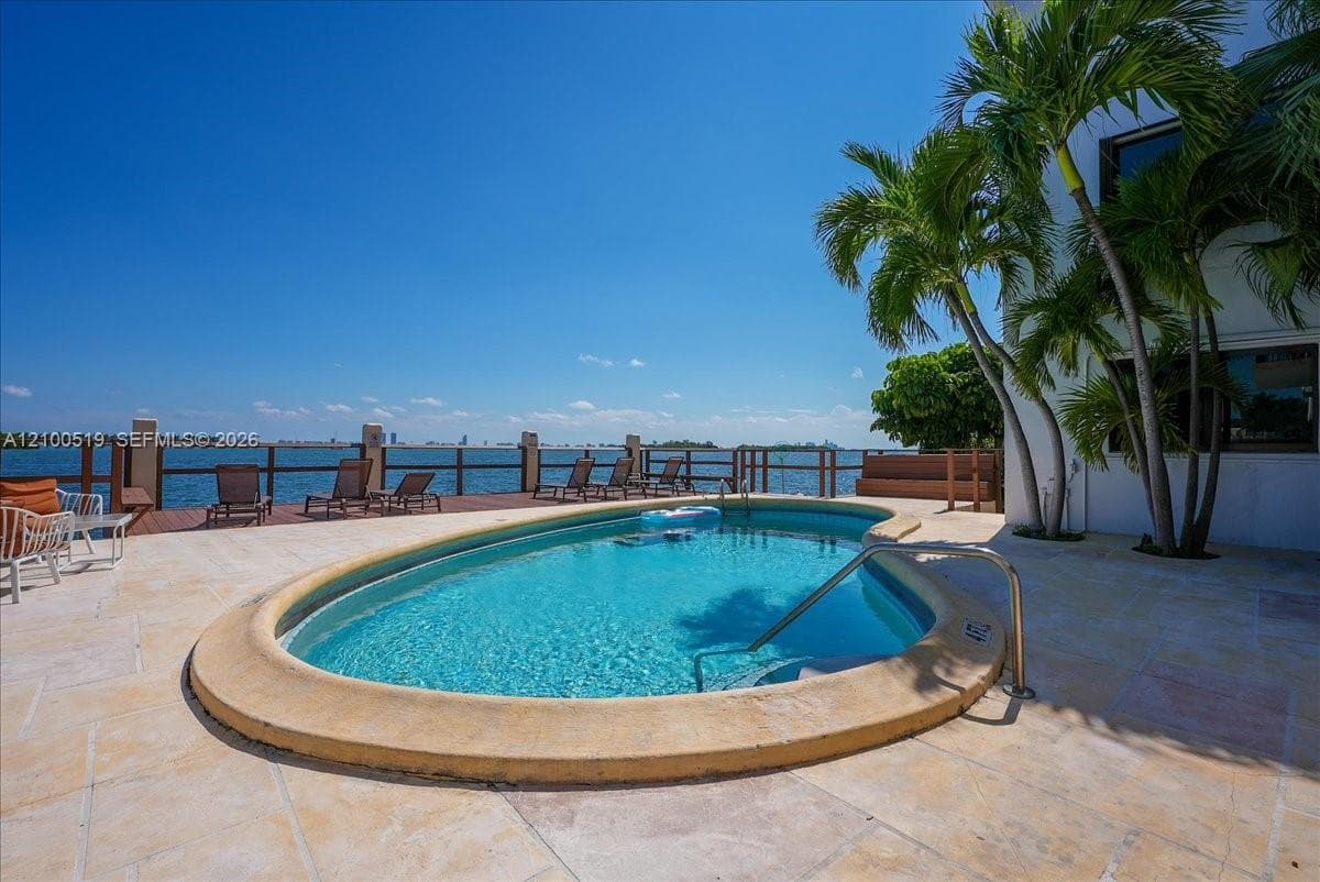 Curved swimming pool with stone coping, expansive stone-paver patio, wooden deck, and waterfront bulkhead
