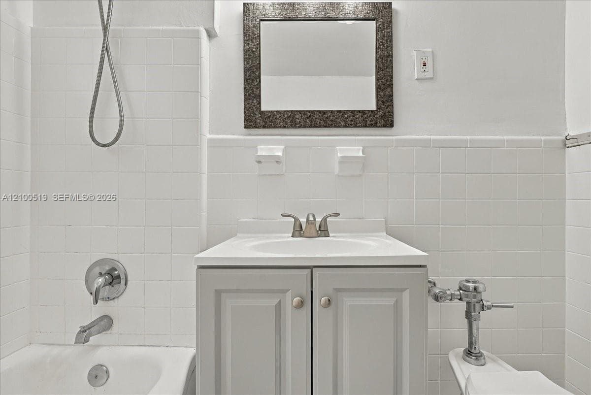 Bathroom featuring white wall tile, grey vanity with integrated sink, chrome dual-handle faucet, wall-mounted mirror with decorative frame, and chrome shower hardware