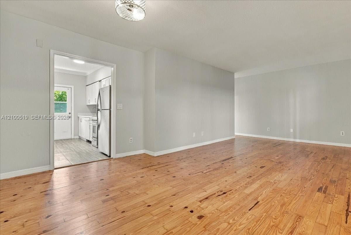 Living area featuring natural wood-finish flooring and a flush-mount drum light fixture, with a view into a kitchen containing white cabinetry and tile flooring