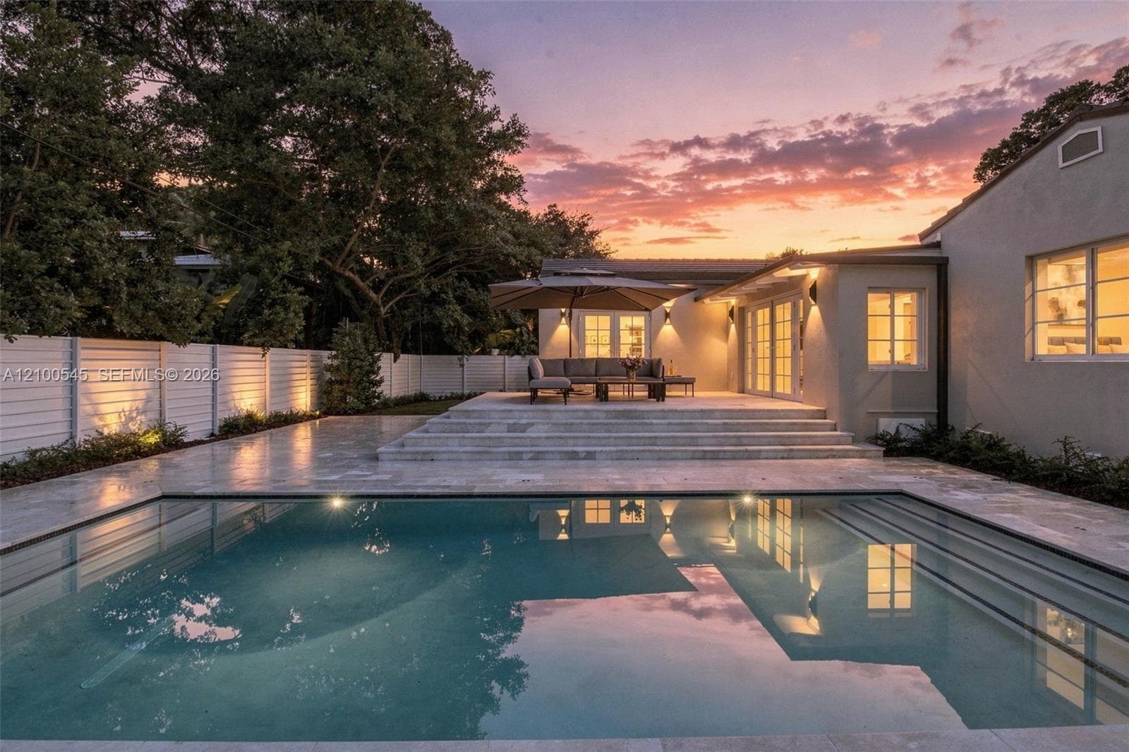 In-ground swimming pool featuring stone-paved decking, multi-tiered steps, a white horizontal-slat perimeter fence, and exterior wall-mounted lighting