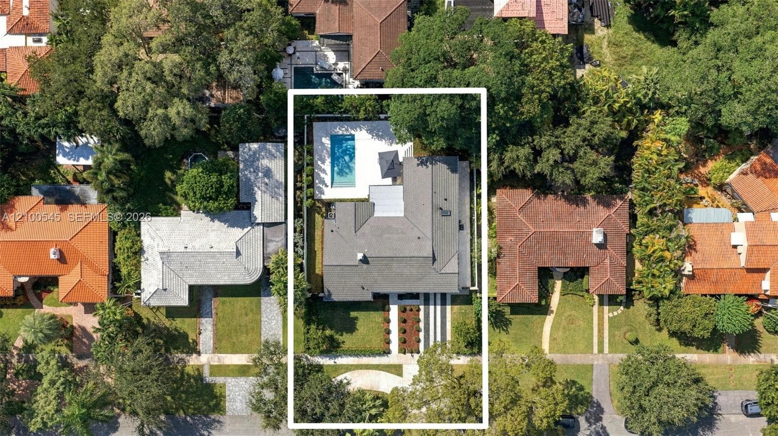 Aerial view of a residential property featuring an in-ground swimming pool, concrete hardscaping, mature canopy trees, and a multi-level roofline