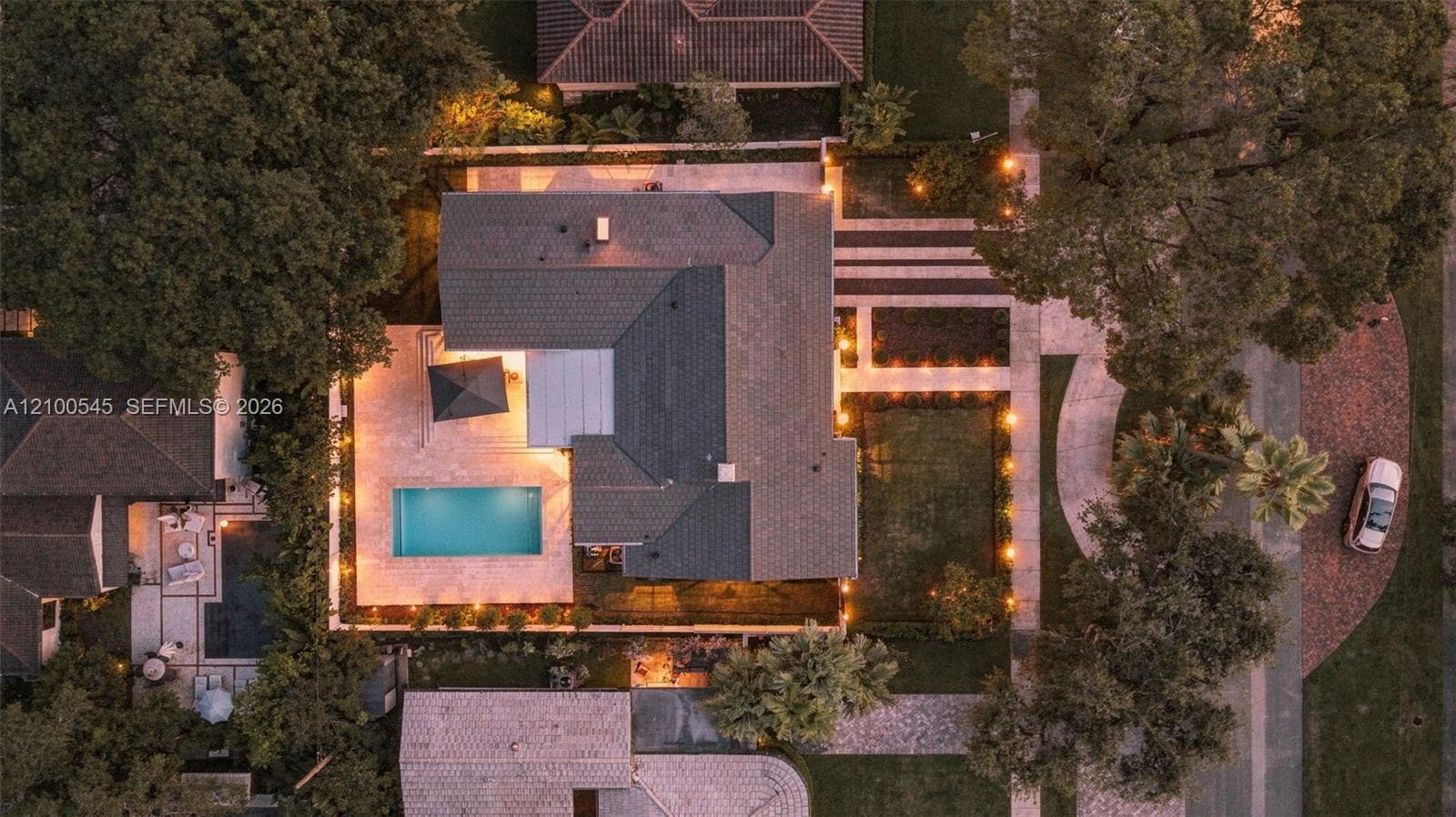 Aerial perspective featuring a rectangular swimming pool, expansive stone-paver hardscaping, mature trees, and structured landscaping with integrated pathway lighting