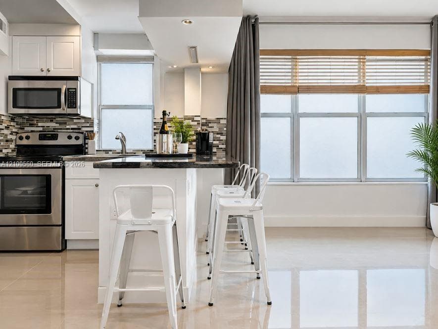 Kitchen featuring white cabinetry, stainless steel appliances, multi-toned tile backsplash, stone-surface breakfast bar, and polished flooring