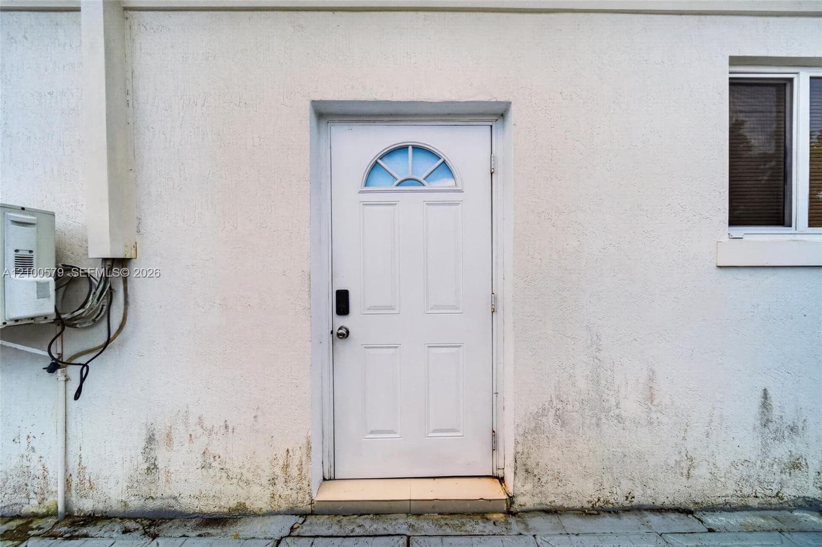 White exterior wall with textured stucco finish, featuring a four-panel entry door with a semi-circular transom window and a rectangular window with a white frame