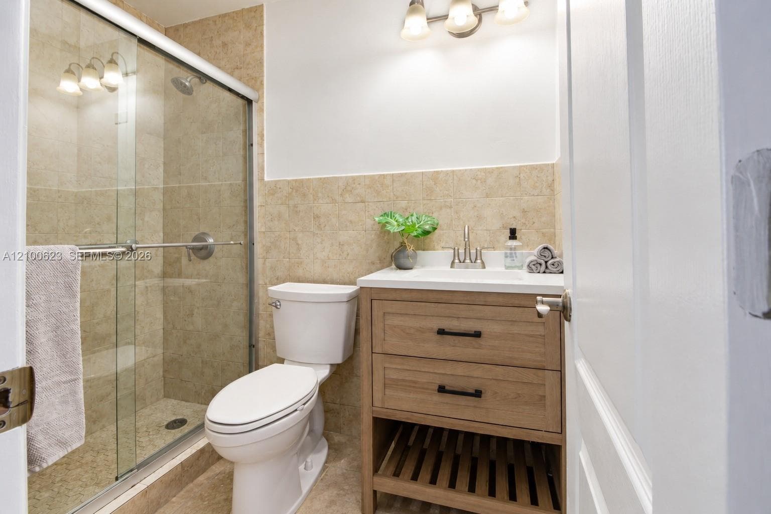Bathroom featuring a wood-finish vanity with two drawers and an open slatted lower shelf, a glass-enclosed shower with tiled surround, and integrated wall-mounted lighting