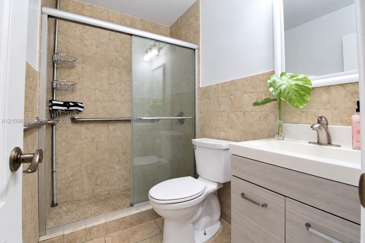 Bathroom featuring a walk-in shower with sliding glass doors, neutral tile surround, integrated metal grab bars, and a wood-finish vanity with a white countertop