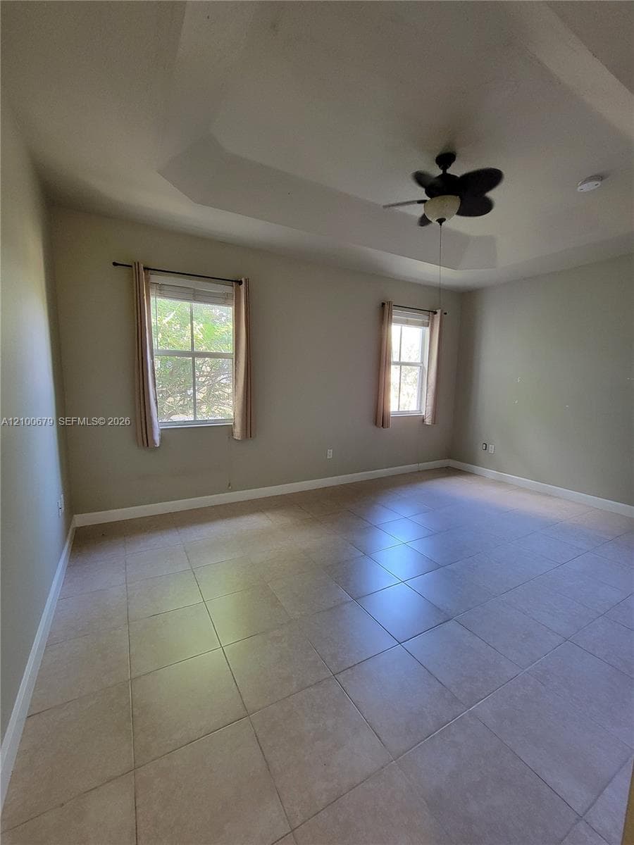 Room featuring a tray ceiling, neutral-toned tile flooring, and a multi-blade ceiling fan