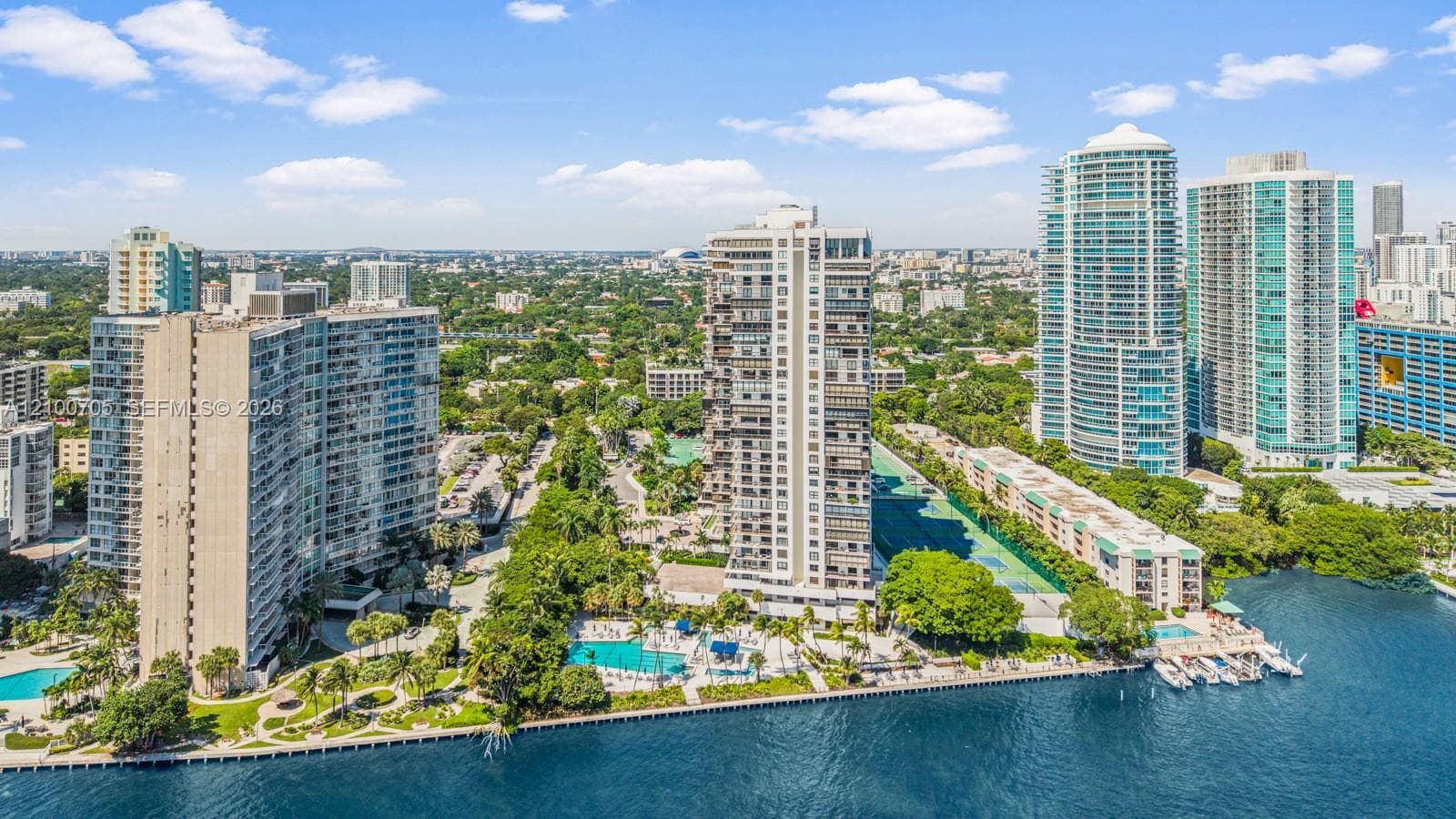 Aerial perspective featuring high-rise architecture, waterfront bulkhead, swimming pools, tennis courts, and landscaped grounds