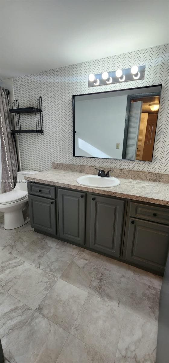 Bathroom vanity featuring dark gray cabinetry, stone-finish countertop with an integrated oval basin, patterned wall covering, and a rectangular mirror with a four-light fixture