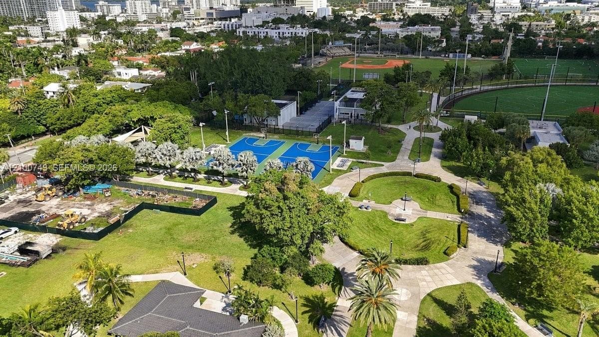 Aerial perspective featuring a multi-story urban skyline, a large athletic field with a synthetic surface and perimeter track, a recreational swimming pool with a water slide, and abundant tropical landscaping