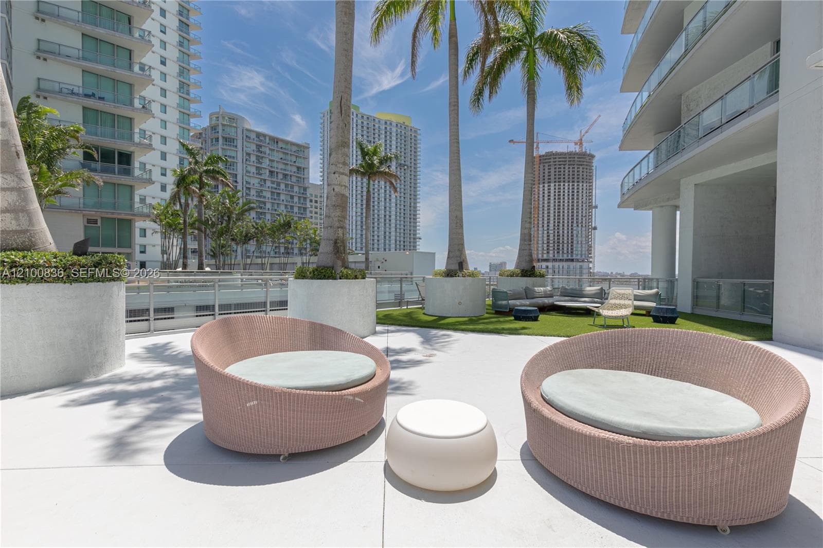 Outdoor terrace featuring a concrete patio surface, manicured synthetic grass area, glass-panel perimeter railing, and decorative cylindrical concrete planters with palm trees