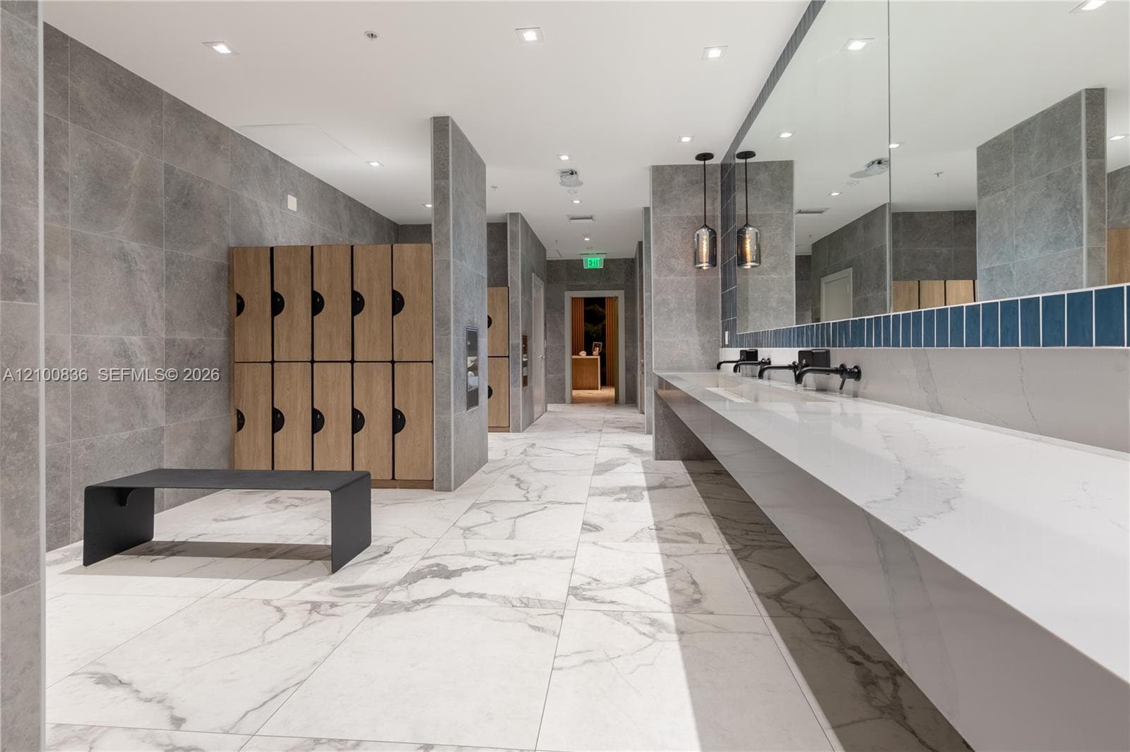 Floating vanity with an integrated countertop, wall-to-wall mirror, wood-finish lockers, floor-to-ceiling gray tile walls, and large-format porcelain tile flooring