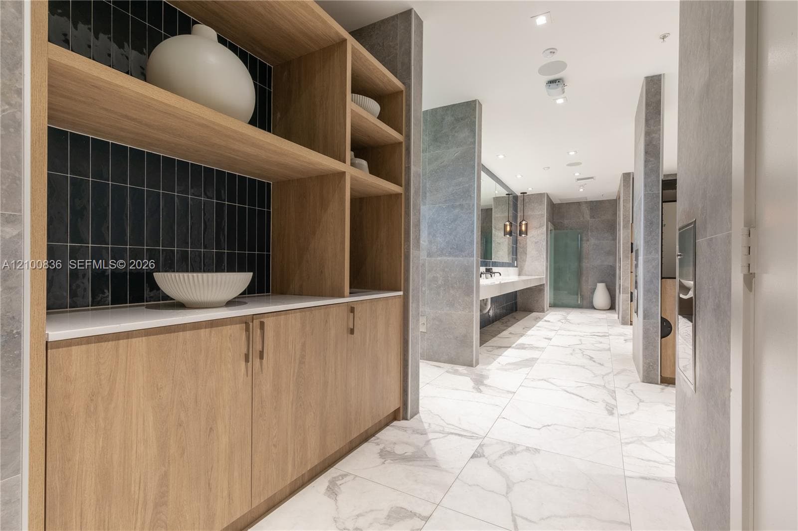 Custom wood-finish cabinetry featuring open shelving and a solid surface countertop with black vertical tile backsplash, paired with large-format stone-finish floor tiling and recessed lighting