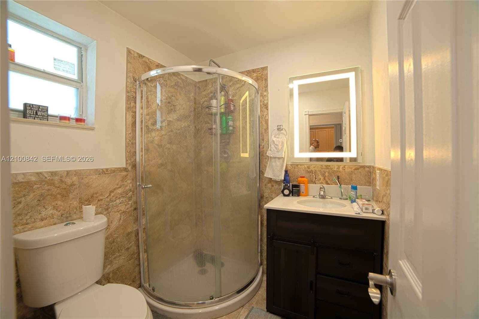 Bathroom featuring a curved glass shower enclosure, stone-finish wall tile, dark-finish wood vanity, integrated surface-mount sink, and rectangular LED-lit mirror