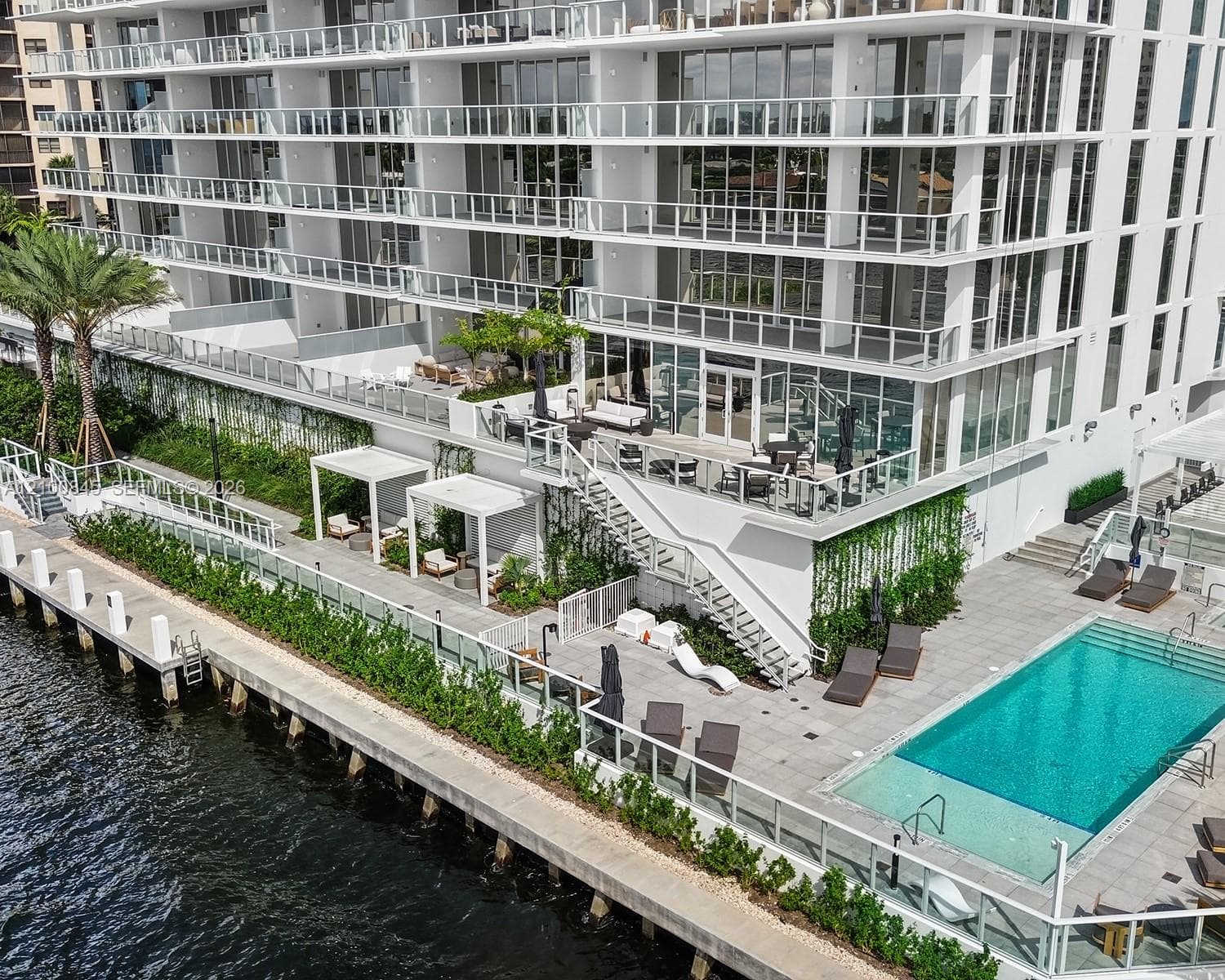 High-rise building exterior featuring tiered balconies with glass railings, a waterfront pool deck, stone-paved sun terrace, integrated pergolas, and vertical landscaping