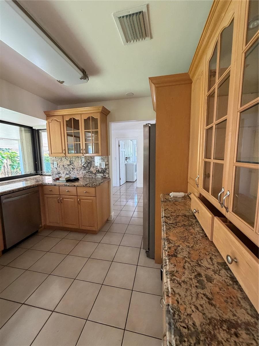 Kitchen featuring light-finish cabinetry with glass-panel upper doors, stone-surface countertops, stone-pattern backsplash, stainless steel dishwasher, and ceramic tile flooring