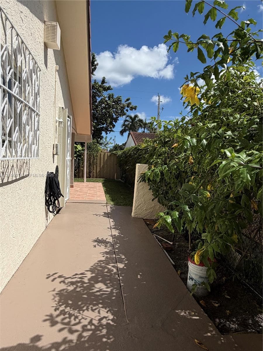 Exterior side yard featuring a concrete walkway, tiled patio area, and white metal security window grilles