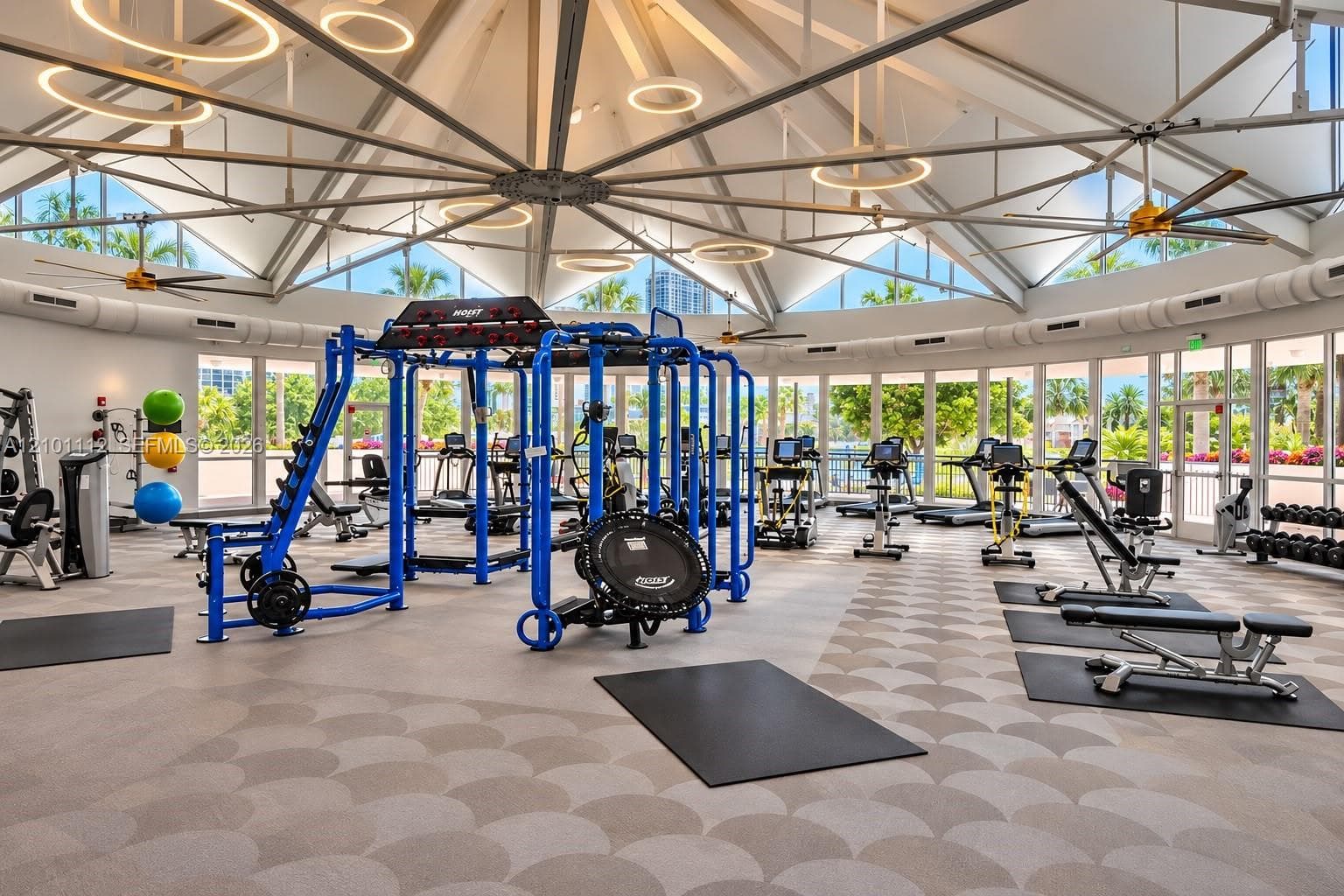 Expansive fitness center featuring a vaulted ceiling with exposed structural beams, circular pendant lighting, full-height glass perimeter walls, and patterned tile flooring