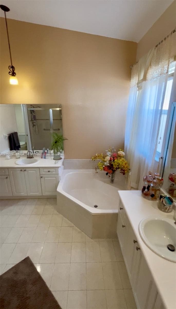 Bathroom featuring dual white vanities with integrated sinks, a built-in corner soaking tub, ceramic tile flooring, and a glass-enclosed shower