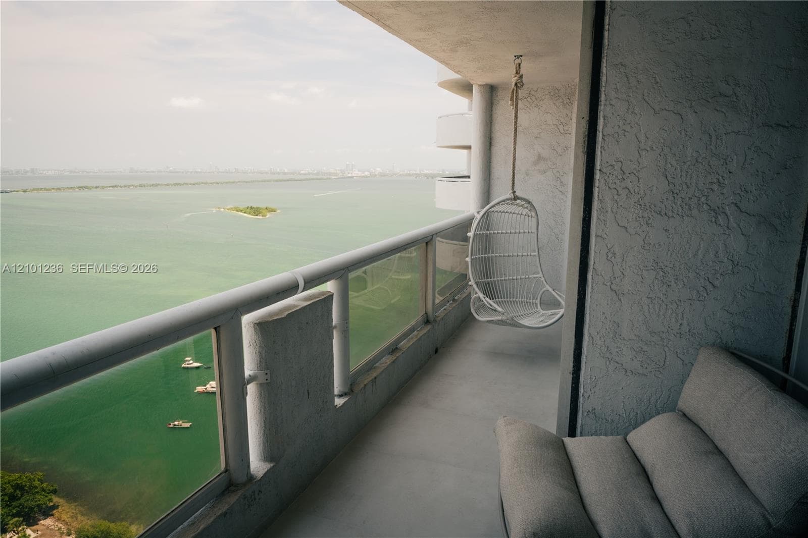 Covered private balcony featuring glass panel railings, textured stucco exterior walls, and water scenery