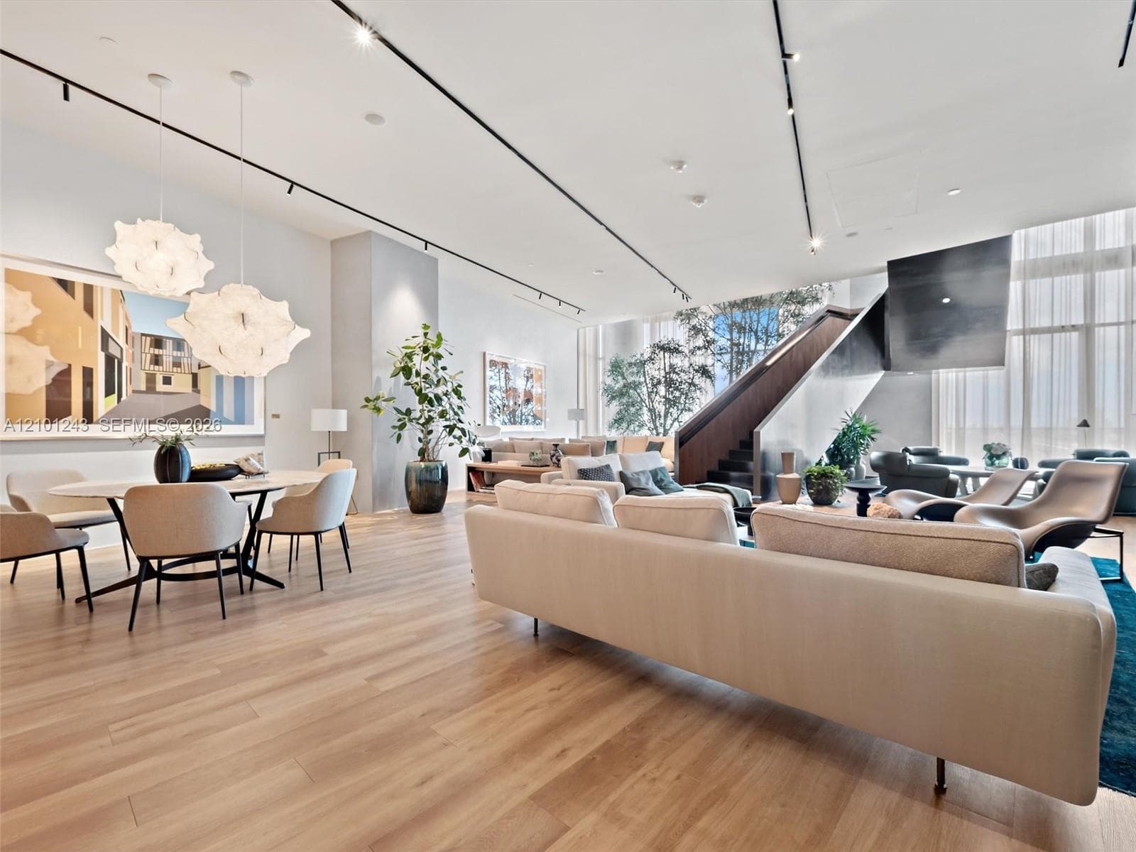 Open-concept main living area featuring light wood-finish flooring, floor-to-ceiling windows, sculptural statement pendant lighting, track lighting, and a floating architectural staircase with a metal railing
