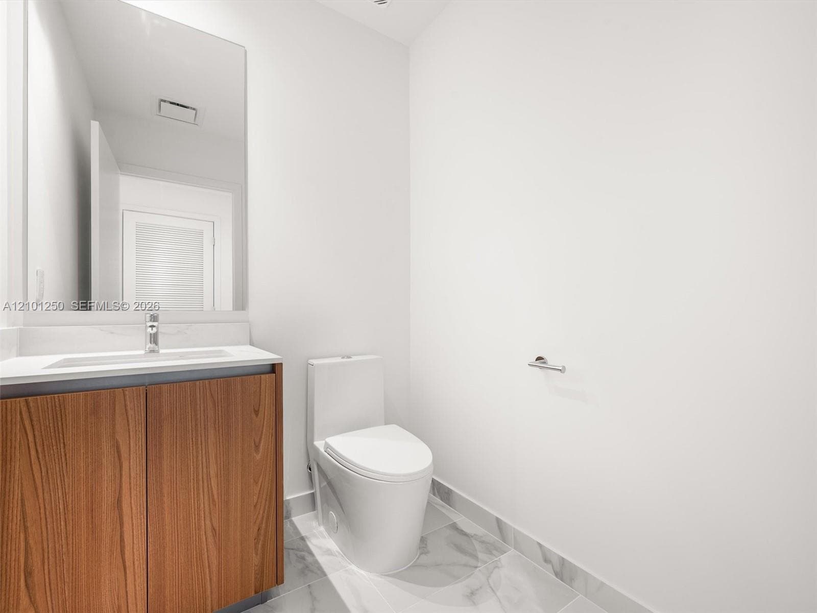 Wood-finish vanity cabinet, solid-surface countertop with integrated sink, chrome faucet, large rectangular wall mirror, and neutral tile flooring