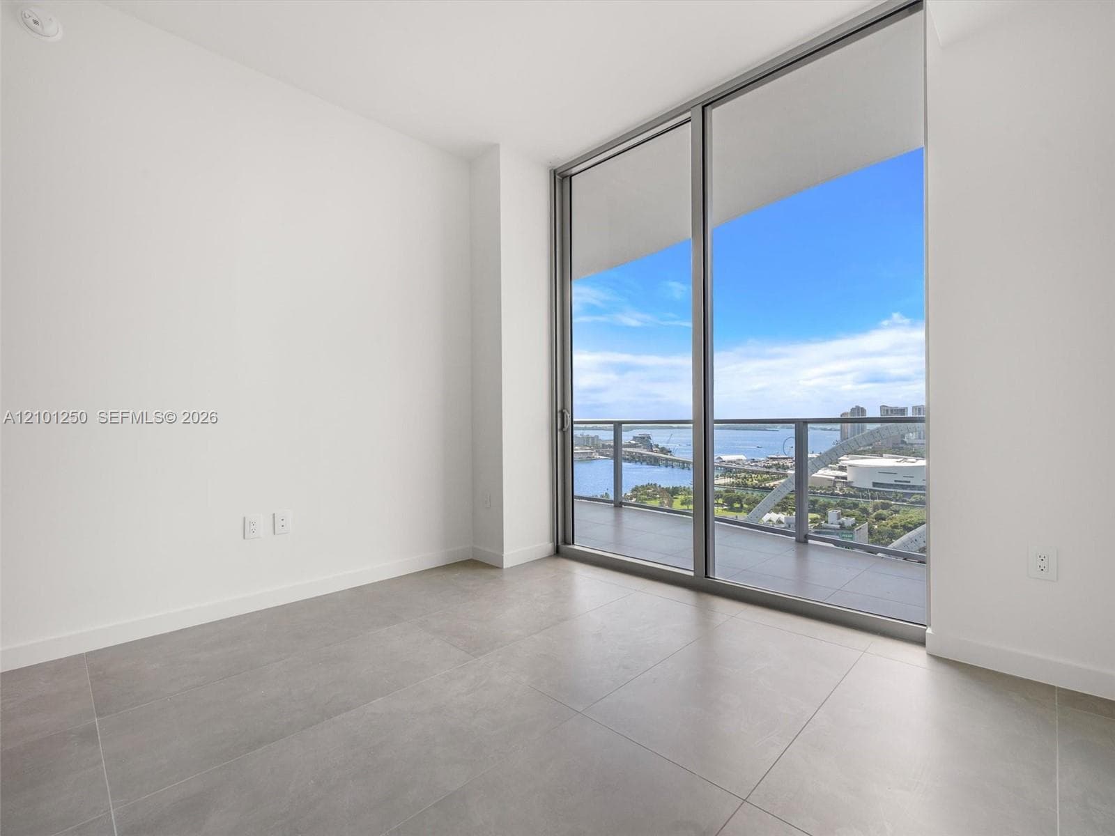 Floor-to-ceiling sliding glass doors, private balcony with metal and glass railing, large-format tile flooring, and water backdrop
