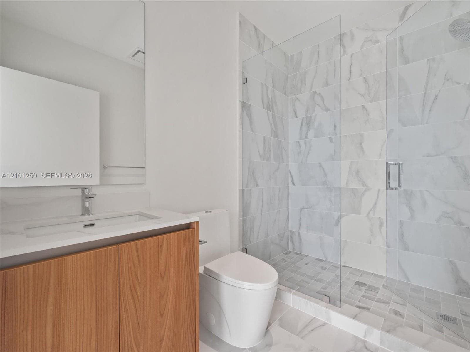 Bathroom featuring a vanity with a light-toned wood-finish cabinet and solid-surface countertop, a frameless glass-enclosed shower with large-format wall tile, and a wall-mounted mirror
