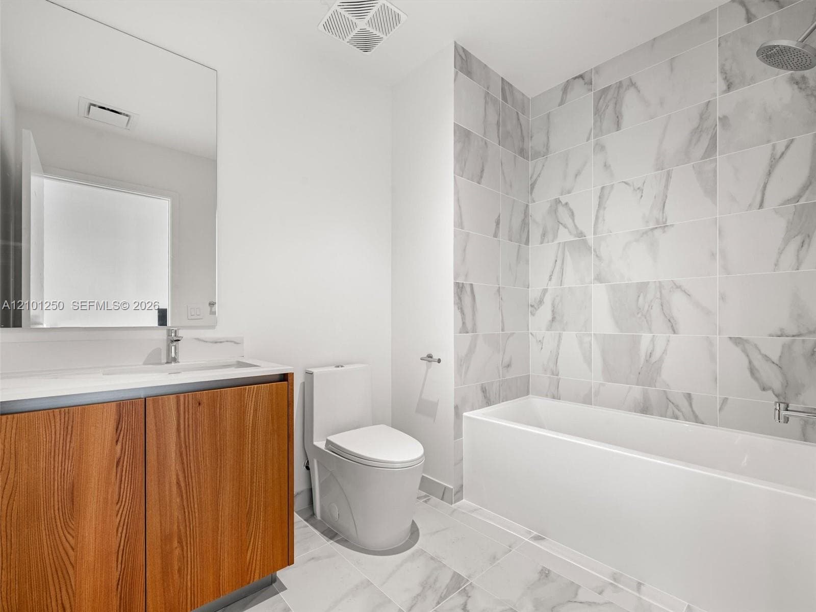 Bathroom featuring a wood-finish vanity with a rectangular sink, large frameless mirror, white plumbing fixtures, and floor-to-ceiling tile surrounding the bathtub
