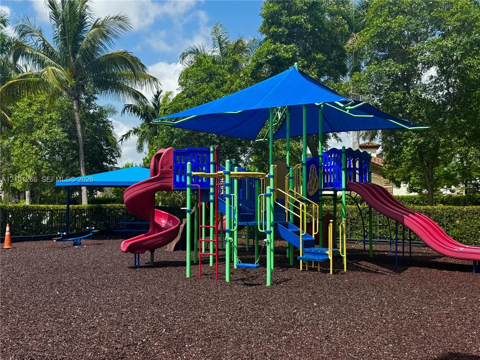Outdoor recreation area featuring a multi-level playset with integrated slides, shade canopy structures, and a rubberized ground surface surrounded by mature landscaping