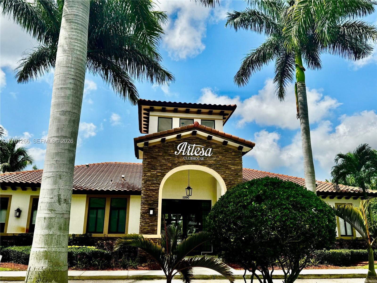 Clubhouse exterior featuring a stone-clad facade, arched entryway, and tile roofing, complemented by mature palm trees and manicured landscaping