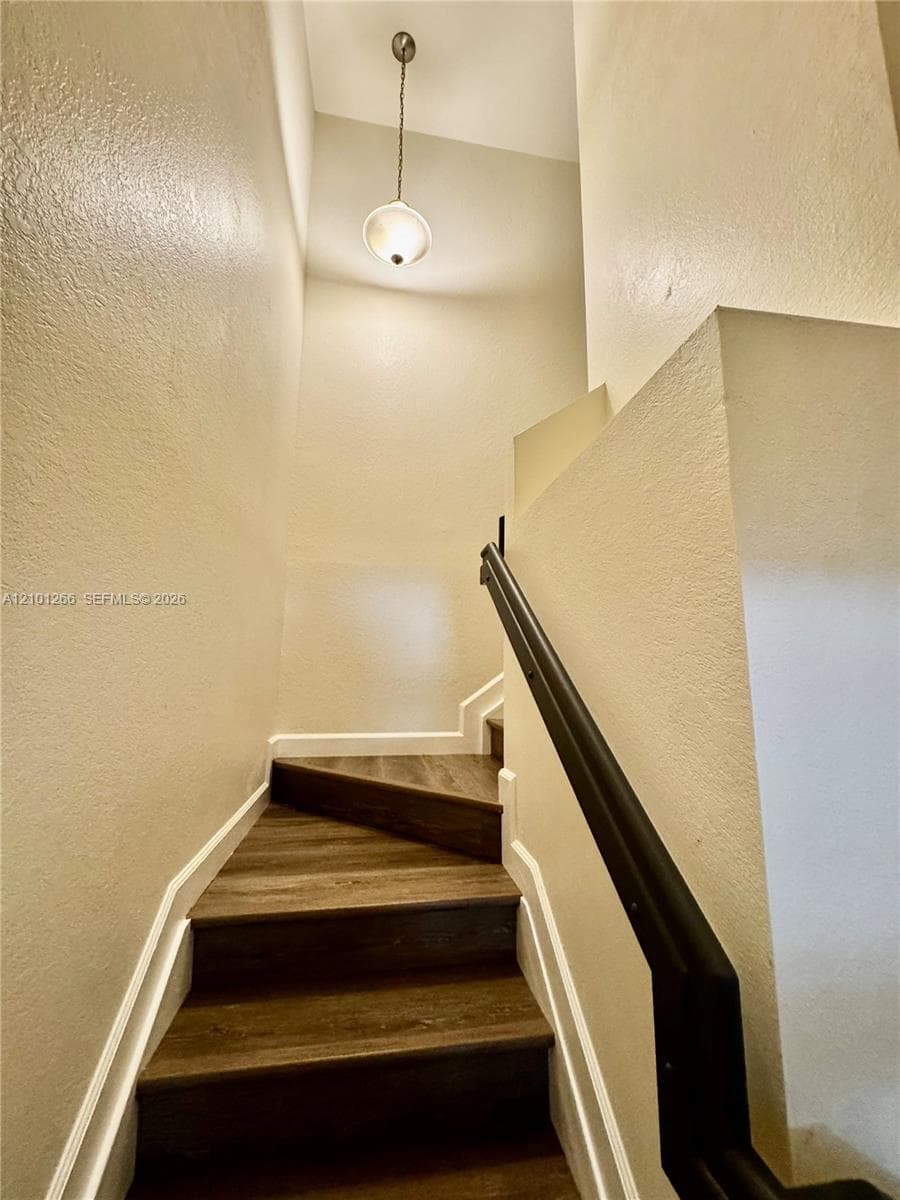 Staircase featuring wood-finish flooring, dark metal handrail, and a suspended globe pendant light